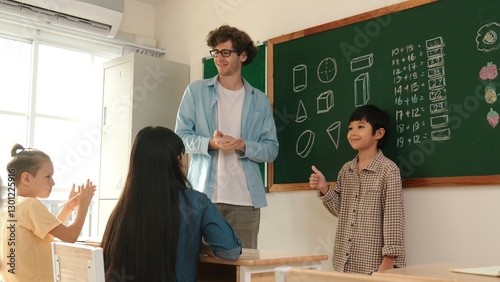 Student standing at in front of class and start to introduce himself at classroom while caucasian teacher looking at student and encourage child. Diverse children sitting and looking at boy. Pedagogy.