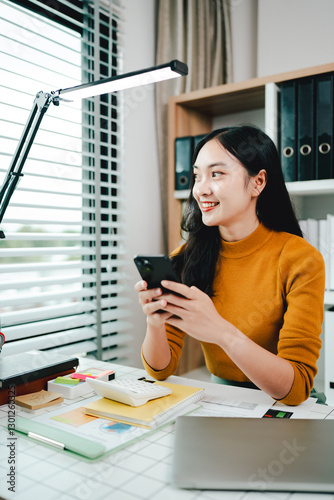 Cheerful beautiful Asian woman uses smartphone to chat at her office. Female accountant makes annual financial and tax reports.