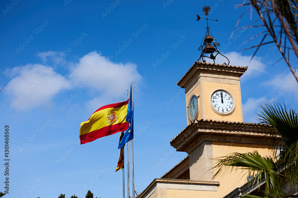 Obraz premium Clock tower with a weathervane and a Spanish flag waving alongside the European Union flag against a blue sky with scattered clouds;