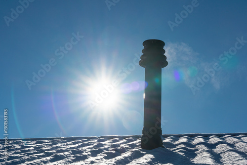 A snow-covered roof with a flue terminal for a gas boiler backlit with the cold winter sun in the background