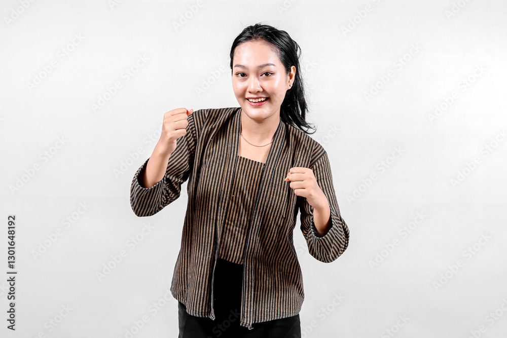 An Indonesian woman wearing traditional lurik attire smiles confidently while raising her fist in a victorious gesture, showcasing cultural pride and enthusiasm against a neutral background. An Indonesian woman wearing traditional lurik attire smiles confidently while raising her fist in a victorious gesture, showcasing cultural pride and enthusiasm against a neutral background.