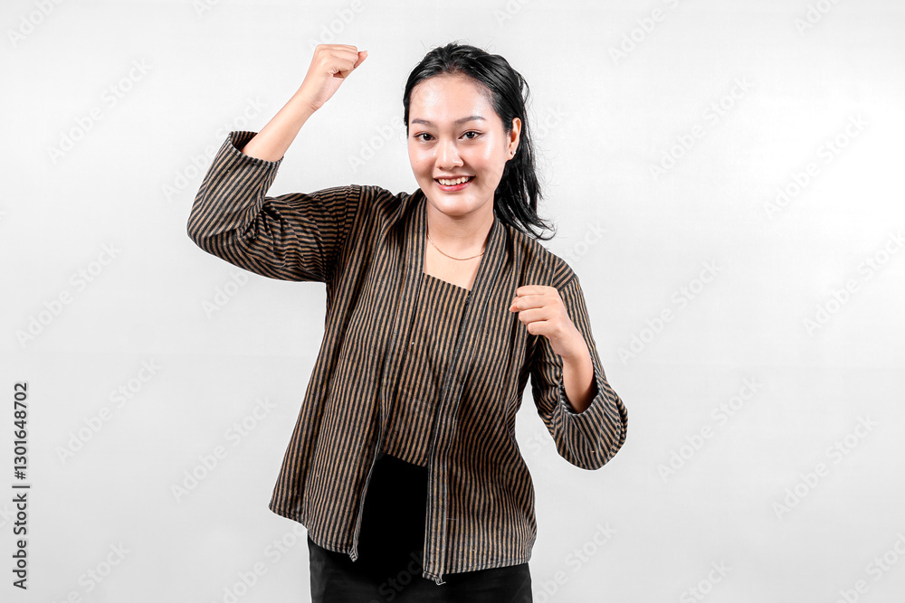 An Indonesian woman wearing traditional lurik attire smiles confidently while raising her fist in a victorious gesture, showcasing cultural pride and enthusiasm against a neutral background. An Indonesian woman wearing traditional lurik attire smiles confidently while raising her fist in a victorious gesture, showcasing cultural pride and enthusiasm against a neutral background.
