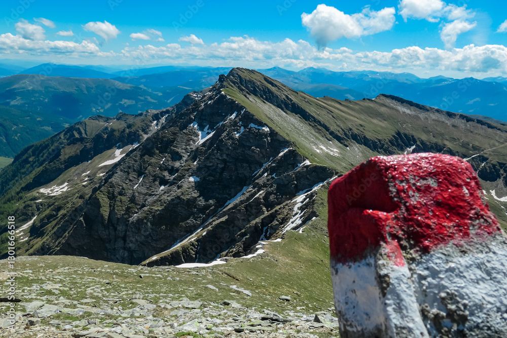 Red and white trail marker stands prominently, directing hikers towards Reitereck peak. Rugged mountains of Ankogel Group rise in the background under a blue, partly cloudy sky in Carinthia, Austria