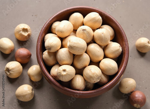 A brown bowl filled with light beige nuts and some scattered around on a neutral background.