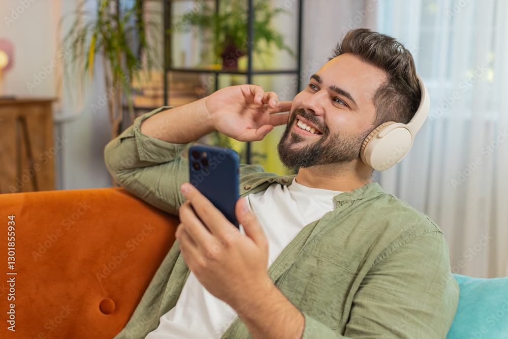 Fototapeta premium Happy overjoyed young man in wireless headphones with smartphone relaxing lying on couch sofa at home apartment choosing, listening favorite energetic disco dancing music. Weekend leisure activities.