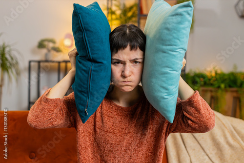 Irritated Caucasian woman girl covers ears with cushions, annoyed by noisy neighbors suffers from headache sitting on couch. Thin walls at home, flat without sound insulation, repair work concept.