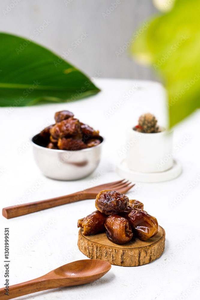 Portrait image of delicious sweet dates, sweet dried dates fruit popular during Ramadan, typical fruit of fasting month. served on white table with ornamental plants, wooden spoon and fork Portrait image of delicious sweet dates, sweet dried dates fruit popular during Ramadan, typical fruit of fasting month. served on white table with ornamental plants, wooden spoon and fork