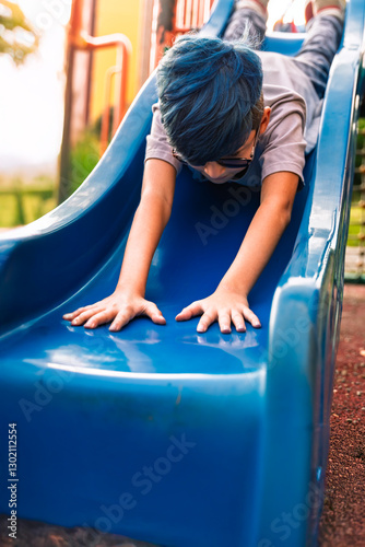 Photo of Colombian Latino mestizo preteen with blue hair sliding down a slide with his head down.