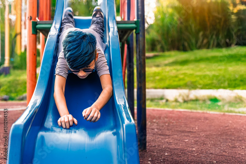 Photo of Colombian Latino mestizo preteen with blue hair sliding down a slide with his head down.