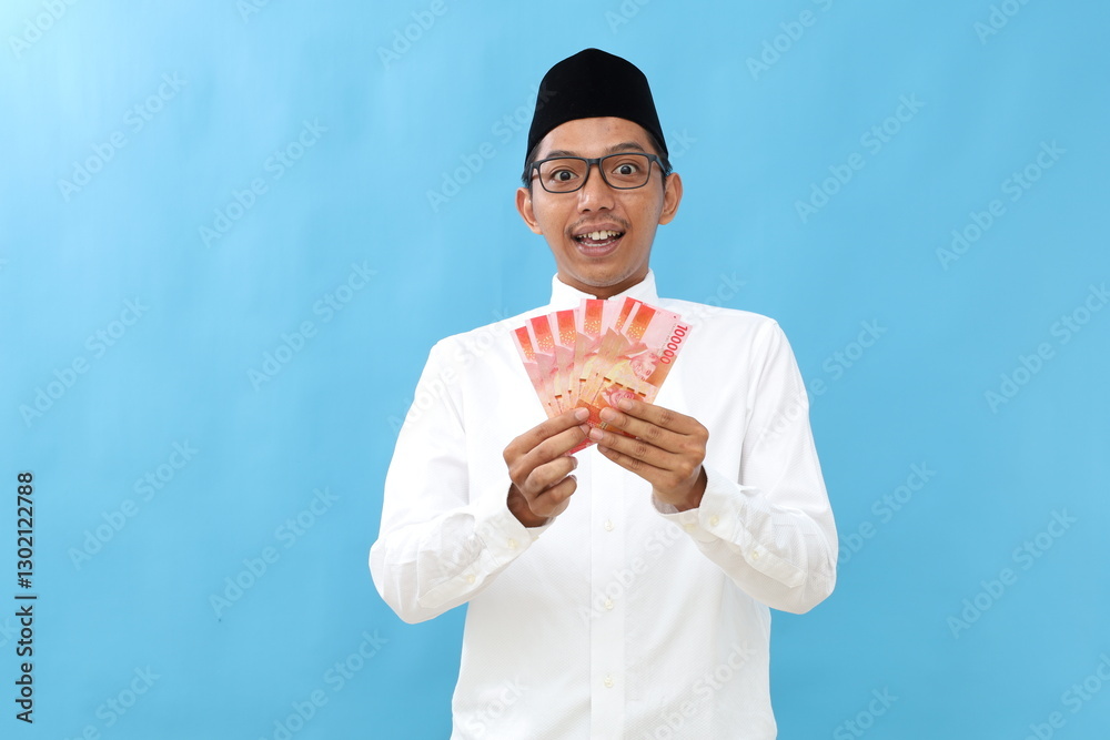Wow excited asian muslim man standing while holding Indonesian banknotes. Isolated on blue Wow excited asian muslim man standing while holding Indonesian banknotes. Isolated on blue