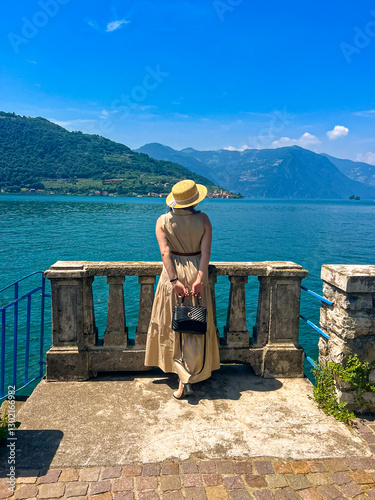 Woman in Maxi Dress and Straw Hat Gazes at a Stunning Mountain-Rimmed Lake Below photo