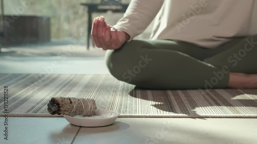 Sunlight illuminates a woman practicing yoga and meditation with her hands in a mudra position, while a smoldering white sage bundle rests on a small plate in front of her
