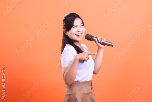 Wallpaper Mural A cheerful young woman holds a microphone, smiling and pointing, ready to perform with passion. isolated on orange background Torontodigital.ca