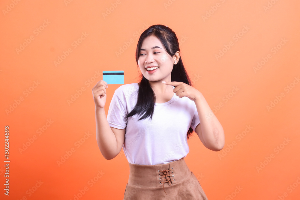 A smiling woman points at a blue credit card, looking pleased and ready to shop, showing off her new financial power, isolated on orange background. A smiling woman points at a blue credit card, looking pleased and ready to shop, showing off her new financial power, isolated on orange background.