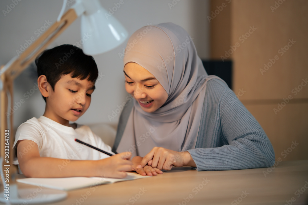Asian Muslim mother wear religious dress and hijab help son with homework at night. Islamic family learn at home. Boy write something on book while woman smile and point. Asian Muslim mother wear religious dress and hijab help son with homework at night. Islamic family learn at home. Boy write something on book while woman smile and point.