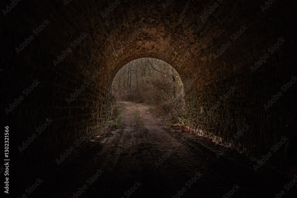 Obraz premium A dark, atmospheric view of a tunnel leading into a brighter, more open space. At the end of the tunnel, an area of ​​brighter light is visible, suggesting an exit or opening into a more open area.