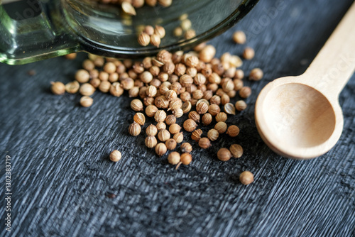 Fototapeta Naklejka Na Ścianę i Meble -  Coriander seeds roll out of a glass jar lying on a dark wooden board. Small wooden measuring spoon