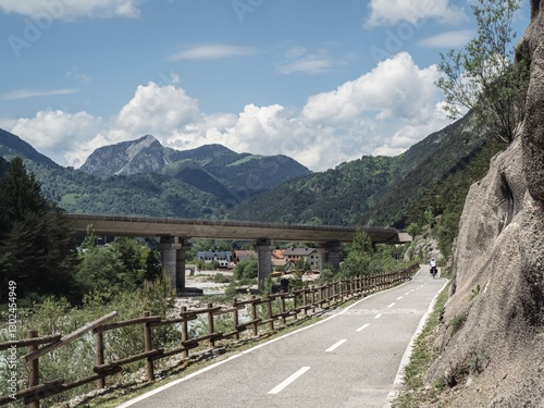 A paved cycling path along the Alpe Adria Radweg in Italy, surrounded by lush green mountains and a scenic river. A bridge spans the valley, while a cyclist enjoys the peaceful landscape.