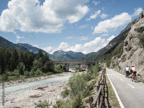 A paved cycling path along the Alpe Adria Radweg in Italy, surrounded by lush green mountains and a scenic river. A bridge spans the valley, while a cyclist enjoys the peaceful landscape.