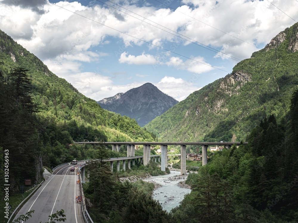 Fototapeta premium A scenic view of a mountain valley in the Italian Alps, featuring a high bridge, a winding road, and a small village nestled among lush green forests under a partly cloudy sky.