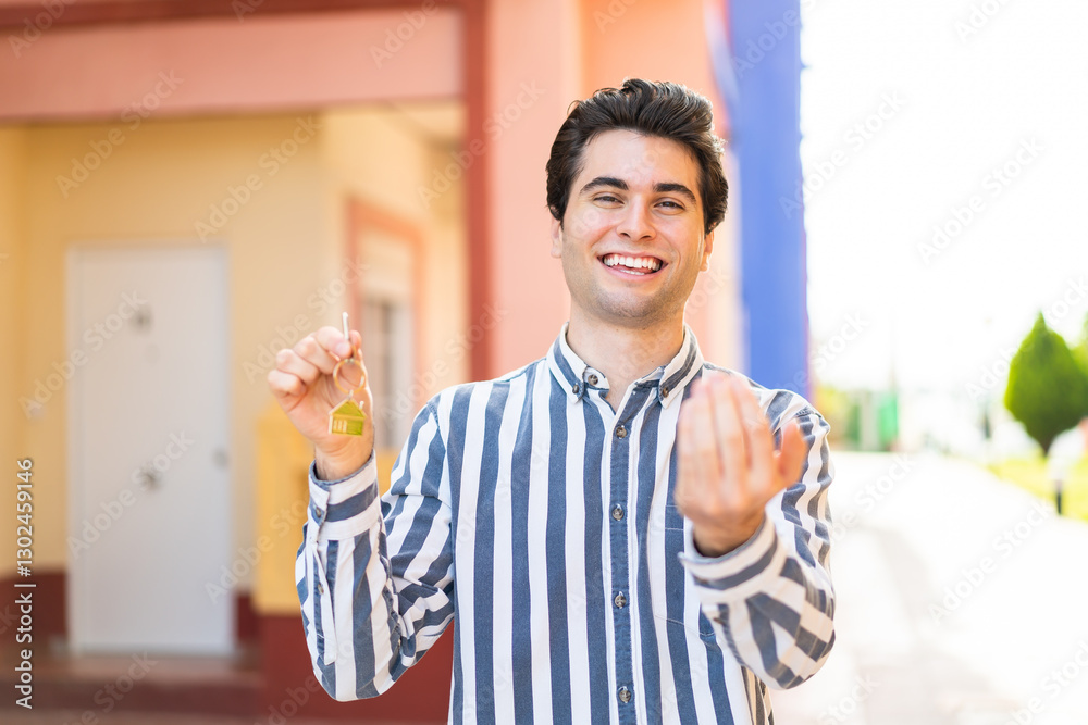 Young handsome man holding home keys at outdoors inviting to come with hand. Happy that you came Young handsome man holding home keys at outdoors inviting to come with hand. Happy that you came