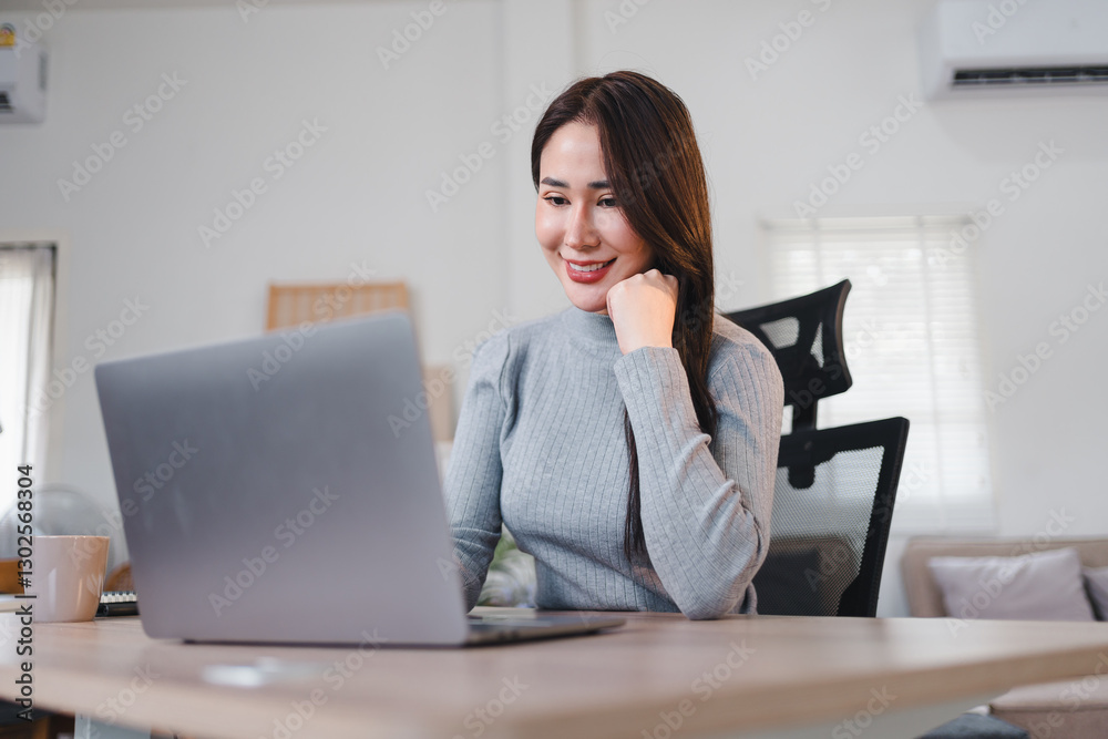 Asian businesswoman in gray sweater working on laptop at desk in bright home office. Asian businesswoman in gray sweater working on laptop at desk in bright home office.