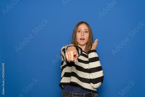 Excited and positive Hispanic teenage girl about 15 years old wearing striped sweater pointing index finger at you and seeing something very funny. Isolated over blue studio background