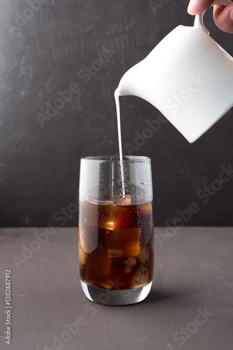 Milk pouring from a milk jug into a glass of iced coffee.