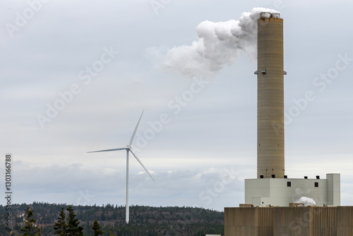 A wind turbine generator behind the smokestack of a fossil fuel generating plant. Overcast sky.