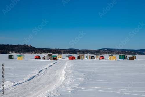 Many ice fishing shacks on a frozen river. Blue sky above. A road in the ice leads to the shacks.