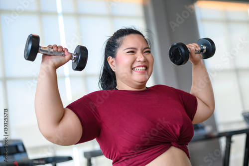 Young, Positive, Smiling, Cheerful overweight woman training with dumbbells in gym. Fat woman in sportswear doing fitness exercise. Weight loss workout, healthy lifestyle concept.