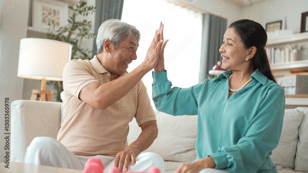 Obraz premium Elder woman using dumbbell at living room for boosting balance and coordination or enhancing joint health and flexibility while grandfather encouraging grandmother and giving high five. Myrmidon.