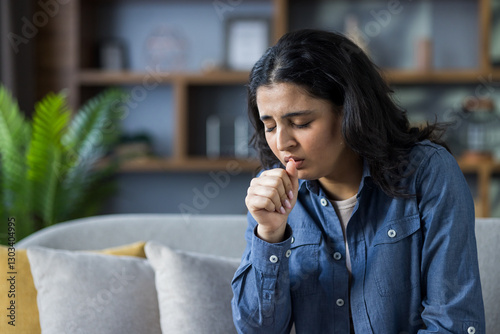 Young woman coughs while sitting on a sofa, possibly experiencing cold or flu symptoms at home.