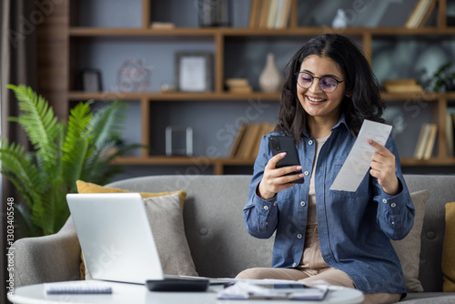Young woman happily manages finances at home using her smartphone for convenient bill payments.