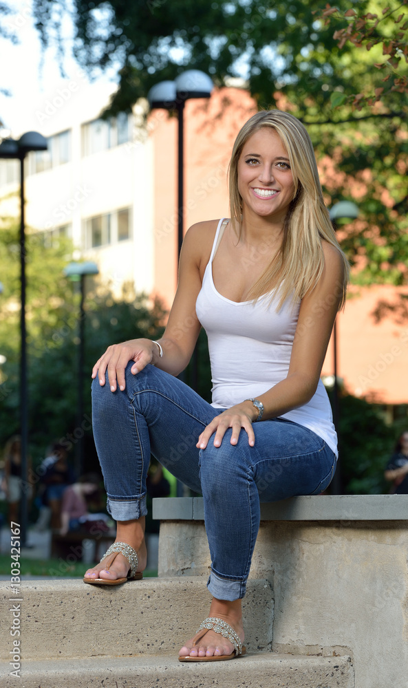 Beautiful young Hispanic woman with blonde hair wearing white tank top and blue jeans - portrait - seated