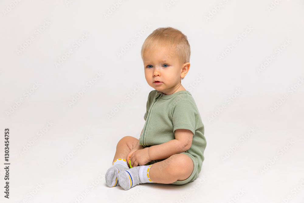 One year little baby boy with blonde hair, in green suit sitting on white background and looking away. Early development concept. Goods and services for children. Copyspace One year little baby boy with blonde hair, in green suit sitting on white background and looking away. Early development concept. Goods and services for children. Copyspace