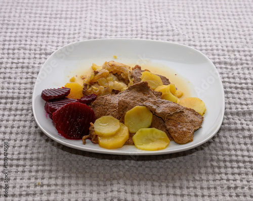 Savory beef slices are accompanied by potatoes and beetrot arranged neatly on a white plate. The hearty meal is set against a soft, textured tablecloth, inviting comfort photo