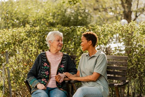 Smiling female caregiver talking with senior woman sitting on bench in park