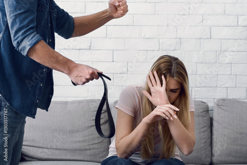 Beating and humiliation. Man threatens with fist and belt to woman, closes face with hands, at home photo