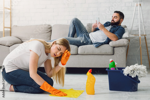 Laziness and indifference in family. Woman in rubber gloves suffers from headache and does cleaning at home, man with beard lies on couch and plays on phone