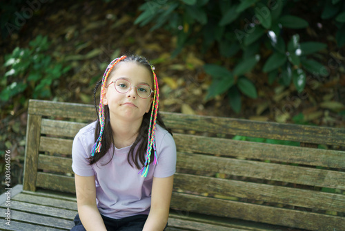 A little sad girl with small colorful braids sitting on a bench during a summer walk in the park. Concept of an active childhood.