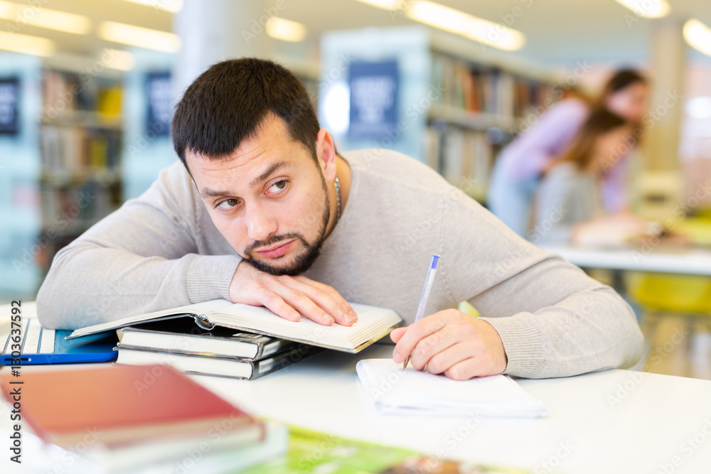 Bored lazy young bearded student sitting at table in university library with his hand and head resting on stack of books.. Bored lazy young bearded student sitting at table in university library with his hand and head resting on stack of books..