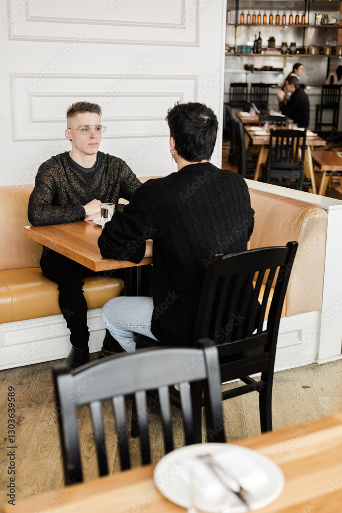 Fototapeta premium A vertical medium shot of a gay men couple on a date in a cozy cafe A mid shot of a LGBTQ+ couple on a romantic date, sitting by a table and talking in an urban cafe interior