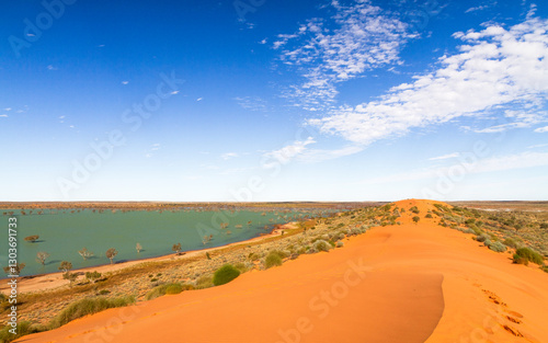 A big red longitudinal sand dune called Big Red on the edge of the Simpson Desert with an unusual ephemeral lake next to it and clouds in a blue sky near Birdsville in Queensland, Australia.