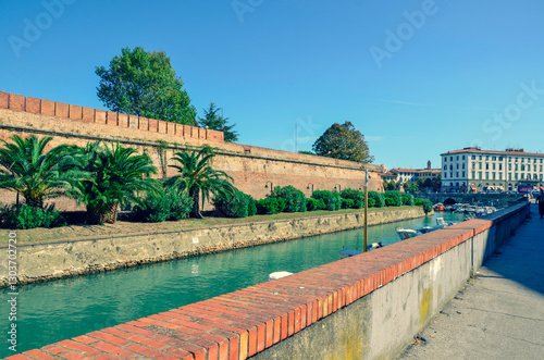 A scenic view of a city canal bordered by the historic walls of a fortress, lined with lush palm trees under a bright, sunny sky. The perspective is from a brick quay.