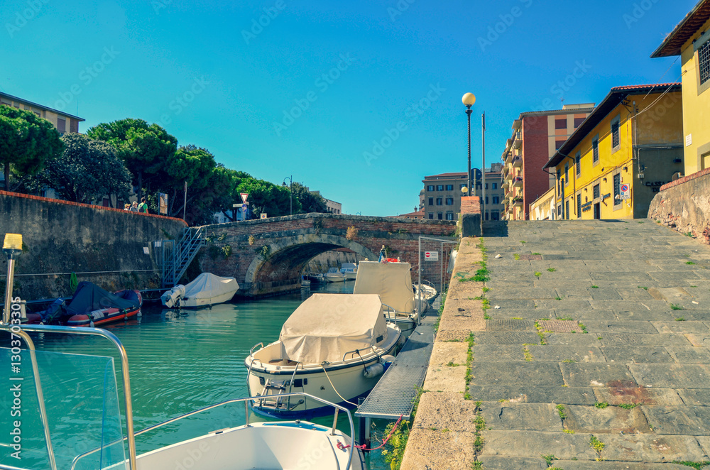 Obraz premium A picturesque city canal on a sunny day. Moored boats line the waterway next to a stone quay and wide steps, with a historic arch bridge visible in the background.
