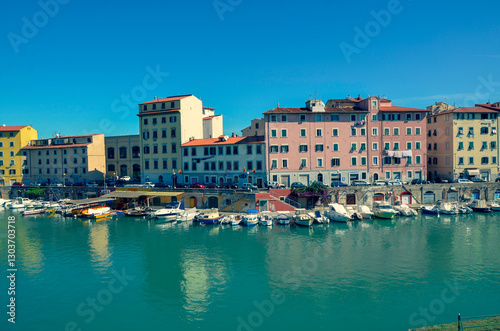Colorful historic buildings line a picturesque city harbor filled with moored boats. The calm turquoise water reflects the vibrant scene under a clear blue sky on a sunny day.