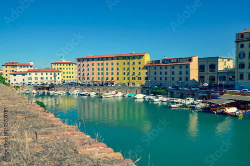 A scenic view from a historic brick wall over a turquoise canal in an Italian city. Colorful buildings and moored boats line the waterway on a bright, sunny day.