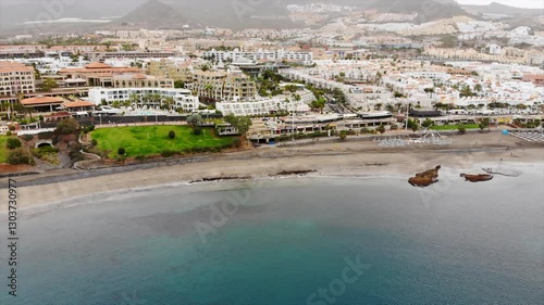 Aerial view of Costa Adeje city, Tenerife, Canary islands. Cinematic 4K footage with sandy coast, calm waters of Atlantic ocean, city modern villas, quay with people, mountains. Early morning video