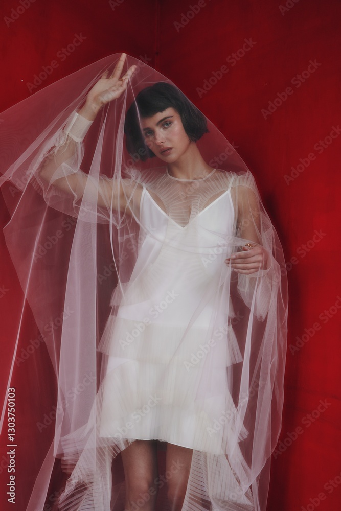 Elegant model poses in sheer fabric dress against a vibrant red background during a fashion photoshoot Elegant model poses in sheer fabric dress against a vibrant red background during a fashion photoshoot